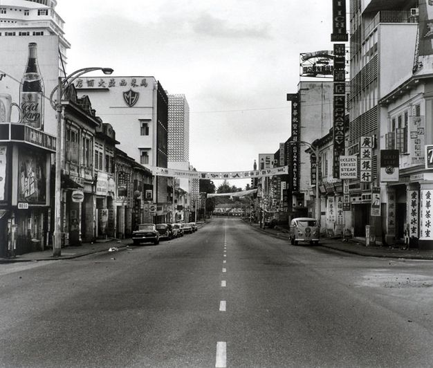 Black and white photograph. An empty two-lane road is lined with buildings hung with signs in Chinese and English. Cars line the curb to our left. A single Volkswagen bus is parked facing us to our right. Two banners are hung from buildings to span the street.