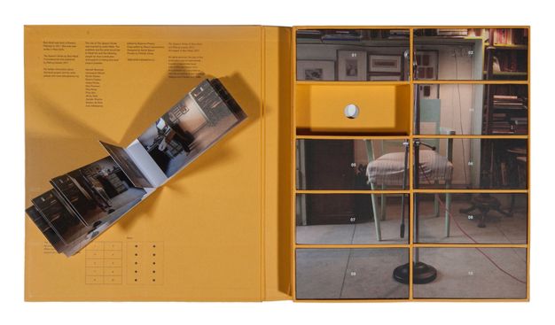 Set of booklets in a case. Ten long books are in a case, two columns across and five rows down, to our right. The second booklet down from the top has been removed and sits open on the cover, which is printed with text.